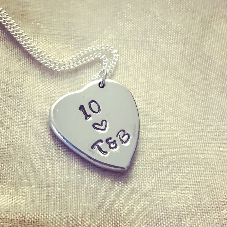 May include: Silver heart-shaped pendant necklace with a chain. The pendant has the text "10" with a heart symbol and "T&B" engraved on it.