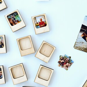Puede incluir: Marcos de fotos de madera con un recorte cuadrado, algunos con fotos coloridas. Los marcos están dispersos sobre una superficie blanca, con fotos sueltas adicionales cerca. Los marcos tienen un acabado de madera natural y un borde oscuro alrededor del recorte.