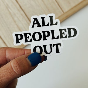 Puede incluir: Una pegatina blanca con el texto "ALL PEOPLED OUT" en letras negras en negrita. La pegatina está sujeta por una mano con una uña pintada de azul. El fondo es una superficie de madera clara.