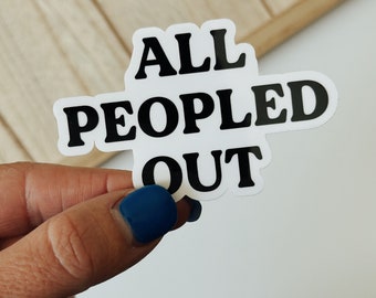 Pode incluir: Um autocolante branco com o texto "ALL PEOPLED OUT" em letras pretas a negrito. O autocolante é segurado por uma mão com uma unha pintada de azul. O fundo é uma superfície de madeira clara.