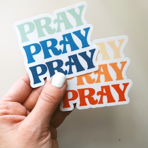 May include: Five colorful stickers with the word "PRAY" in a retro font. The stickers are stacked on top of each other, with the top sticker being light blue, the second sticker being blue, the third sticker being orange, the fourth sticker being yellow, and the bottom sticker being red.
