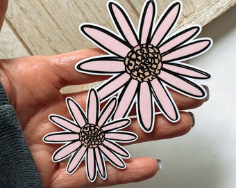May include: Two pink daisy stickers with black outlines. The larger daisy has a brown center and the smaller daisy has a light brown center.