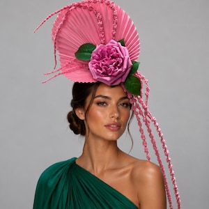 May include: A decorative headpiece featuring a large, circular, pleated pink fan base adorned with a prominent mauve rose and green leaves. Long, thin, textured pink strands extend from the fan, cascading downwards and outwards in a curved shape.
