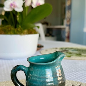 May include: A small, handcrafted turquoise ceramic creamer with a dark brown base and a handle. The creamer is on a decorative mat with a floral design. A white orchid plant is in the background.