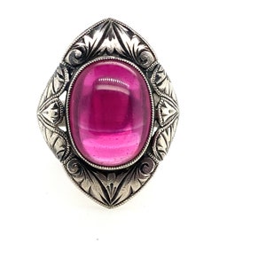 May include: A silver-toned ring with an oval, magenta-colored stone. The ring has an ornate, engraved design around the stone, featuring floral and scroll patterns. The setting is a detailed, vintage-style design.