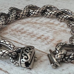 May include: A silver bracelet with a snake-like chain design and a decorative clasp. The bracelet is made of metal and has a textured surface.