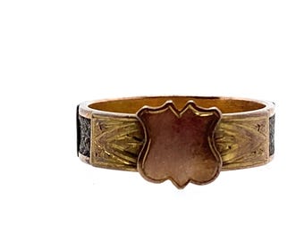 Puede incluir: Un anillo de oro con un diseño en forma de escudo. El escudo está grabado con un patrón floral y tiene un espacio en blanco en el centro. El anillo está engastado con esmalte negro.