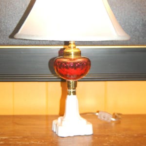 May include: A table lamp featuring a white lampshade and a red glass base. The lamp has a brass stem and a small red orb at the top. The base is white with a geometric design. The lamp is on a wooden surface. The lamp is approximately 40 cm tall.