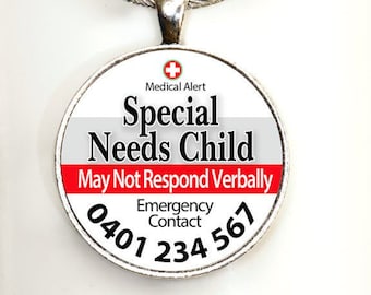 Puede incluir: Un llavero plateado con una etiqueta redonda blanca que dice "Alerta médica Niño con necesidades especiales Puede que no responda verbalmente Contacto de emergencia 0401 234 567". La etiqueta tiene una cruz roja y texto rojo.