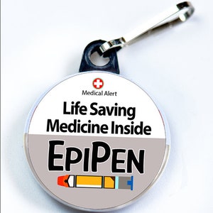 Op de afbeelding: Een ronde medische alarmtag met de tekst "Life Saving Medicine Inside" en "EpiPen". De tag heeft een rood kruissymbool en een karabijnhaak. De EpiPen is afgebeeld in oranje, geel en blauw.