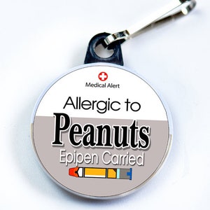 Può includere: Un tirare cerniera rotondo, bianco e grigio con una croce rossa e il testo "Medical Alert" in nero. Il testo "Allergic to Peanuts" è in nero con il testo "EpiPen Carried" in nero sotto. Un'immagine di cartone animato di un EpiPen è sotto il testo.