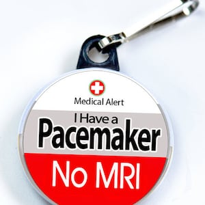May include: A round medical alert tag with a silver clip. The tag is white, gray, and red, with the words "Medical Alert" and "I Have a Pacemaker No MRI" in black text. A red cross symbol is at the top.
