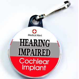 May include: A round, white, grey, and red zipper pull with the text "Medical Alert", "HEARING IMPAIRED", and "Cochlear Implant".