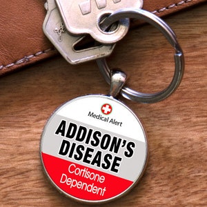 Puede incluir: Un llavero plateado con una etiqueta redonda de alerta médica. La etiqueta es roja, blanca y gris con el texto "Alerta médica", "Enfermedad de Addison" y "Dependiente de la cortisona".