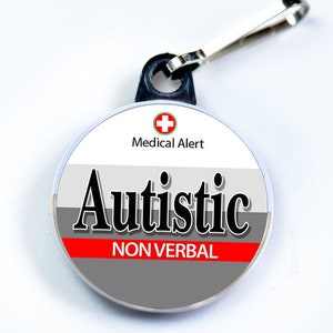 Puede incluir: Una etiqueta de alerta médica redonda y blanca con una cruz roja y el texto "Medical Alert". La etiqueta también tiene una franja gris con la palabra "Autistic" en negro y una franja roja con el texto "NON VERBAL" en negro.