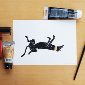 May include: A black and white linocut print of a dachshund dog lying on its back with a yellow collar. A tube of black paint, a tube of gold paint, a paint roller, and a paintbrush are also visible on a wooden surface.