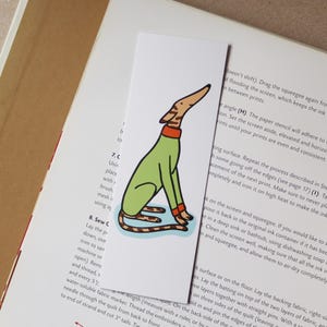 May include: A whimsical bookmark featuring a cartoon dog illustration. The dog is depicted in a seated position, wearing a green outfit with orange accents. The bookmark is rectangular and placed on a book page.