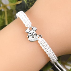 Könnte beinhalten: Weißes Makramee-Armband mit einem silberfarbenen Mäuse-Charm. Das Armband ist mit Gleitknoten verstellbar. Der Charm hat ein detailliertes Mäuse-Design. Das Armband wird am Handgelenk gezeigt.