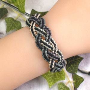 以下が含まれることがあります： 手首につけられた黒、白、緑の編まれたブレスレット。ブレスレットは編まれたコードで作られており、シンプルでミニマルなデザインです。