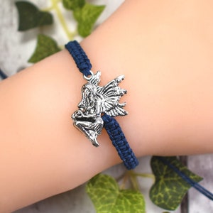 May include: A navy blue braided bracelet with a silver fairy charm. The bracelet is tied on a person's wrist.