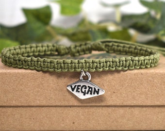 May include: A green braided bracelet with a silver charm that says "VEGAN". The bracelet is on a brown box.
