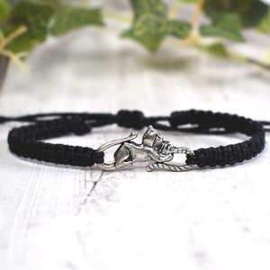 May include: A black braided bracelet with a silver cat charm. The charm is of a cat sitting on its hind legs with its tail curled around its body.