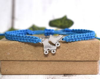 May include: A blue macrame bracelet with a silver roller skate charm. The bracelet is displayed on a small brown cardboard box. The background is a blurred white surface with green foliage.