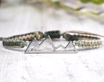 May include: A braided bracelet with a silver mountain charm. The bracelet is made with green, brown, and beige cord.
