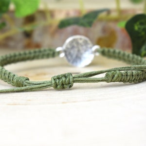 May include: A green braided bracelet with a silver circular charm. The bracelet is tied with a knot.