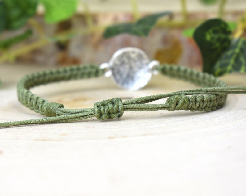 May include: A green braided bracelet with a silver circular charm. The bracelet is tied with a knot.