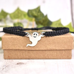 Puede incluir: Pulsera de cordón trenzado negro con un dije de fantasma plateado. La pulsera está sobre una caja marrón.