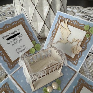May include: A handmade baby announcement card with a light blue and brown design. The card features a small wooden crib, a stork carrying a baby, and the baby's birth details: Enzo Angelino, June 28th, 2025, 7 lbs 5 oz, 20 inches.