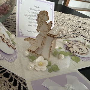 May include: A handmade, multi-layered greeting card with a religious theme. The card features a wooden cutout of a girl praying, surrounded by decorative elements like pearls, flowers, and a cross. The card is white and lavender.