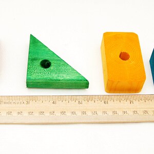 Puede incluir: Cuatro bloques de madera de diferentes formas y colores: un prisma rectangular rojo, un tri&aacute;ngulo verde, un prisma rectangular amarillo y un cubo azul. Cada bloque tiene un agujero perforado en el centro. Los bloques est&aacute;n dispuestos en una fila sobre una superficie blanca con una regla debajo.
