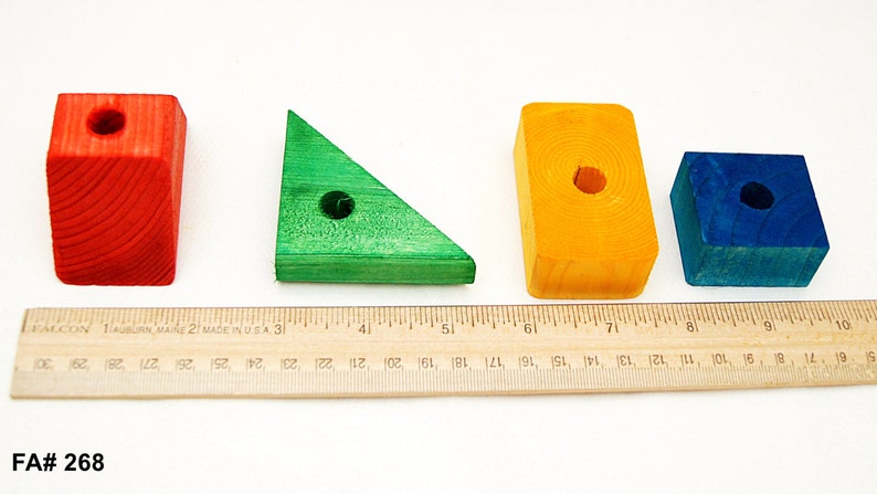 Puede incluir: Cuatro bloques de madera de diferentes formas y colores: un prisma rectangular rojo, un tri&aacute;ngulo verde, un prisma rectangular amarillo y un cubo azul. Cada bloque tiene un agujero perforado en el centro. Los bloques est&aacute;n dispuestos en una fila sobre una superficie blanca con una regla debajo.