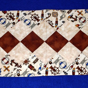 May include: A beige and brown table runner with a nautical theme. The runner features a repeating pattern of anchors, ships' wheels, ropes, and life preservers. The runner is made of fabric and has a diamond pattern.