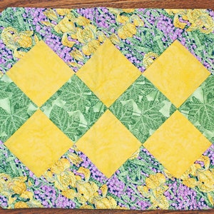 May include: A yellow and green quilted table runner with a floral pattern. The runner is made of fabric squares sewn together in a diamond pattern.