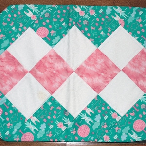 May include: A decorative table runner with a diamond pattern. The centre features alternating pink and white squares. The border is teal with a repeating pattern of bunnies, trees, and other shapes. The runner is approximately 61 cm long.