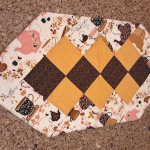 May include: A quilted table runner with a tea party theme. The runner features a central pattern of alternating brown and yellow diamonds. The edges are decorated with a white fabric printed with teacups, teapots, and floral designs.