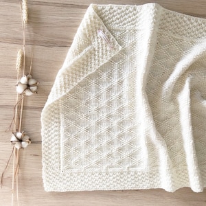 Könnte beinhalten: Eine weiße gestrickte Babydecke mit einem Rautenmuster. Die Decke ist gefaltet und auf einer Holzoberfläche mit Baumwollknäueln und Weizenhalmen gelegt.