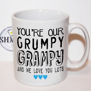 May include: White ceramic mug with black text that reads "You're our grumpy grampy and we love you lots". There are three blue hearts at the bottom of the mug.