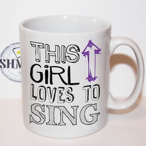 Op de afbeelding: Witte keramische mok met de tekst "THIS GIRL LOVES TO SING" in zwart en paars. De mok heeft een handvat en een eenvoudig ontwerp, perfect voor een muziekliefhebber.