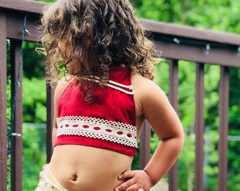 May include: A young girl wearing a red halter top with white lace trim and a white and gold patterned skirt with a red trim. The girl is standing on a wooden deck.