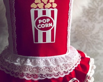 May include: Red and white fabric apron with a popcorn design. The apron has a white lace trim and white pom-poms around the bottom.