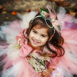 Pode incluir: Uma menina pequena usando um vestido de tule rosa e branco com uma faixa de cabeça floral. Ela está sorrindo e olhando para a câmera.