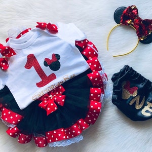 Può includere: Un tutù nero e rosso con bordo in pizzo bianco, un body bianco con un numero uno in glitter rosso e una testa di Topolino nera, un pannolino in raso nero con scritta in glitter oro che dice "uno" e una fascia per capelli dorata con un fiocco di paillettes nero e rosso.