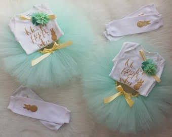 May include: Two baby girl outfits for Easter. Each outfit includes a white bodysuit with gold glitter text that says "Oh... for peep's sake", a green tulle tutu, a yellow ribbon, and white leg warmers with a gold glitter bunny.