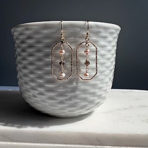 Könnte beinhalten: Goldene Ohrringe mit einem zarten, strukturierten ovalen Rahmen und einer herabhängenden Kette aus rosa Perlen und klaren Kristallen.