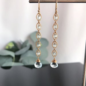 Op de afbeelding: Gouden oorbellen met een delicaat kettingontwerp en lichtblauwe edelsteenaccenten. De oorbellen hebben een traanvormige edelsteen aan de onderkant.