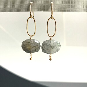 K&ouml;nnte beinhalten: Goldene Ohrringe mit einem grauen und schwarzen Edelstein-Anh&auml;nger. Die Ohrringe haben ein schlichtes ovales Design.